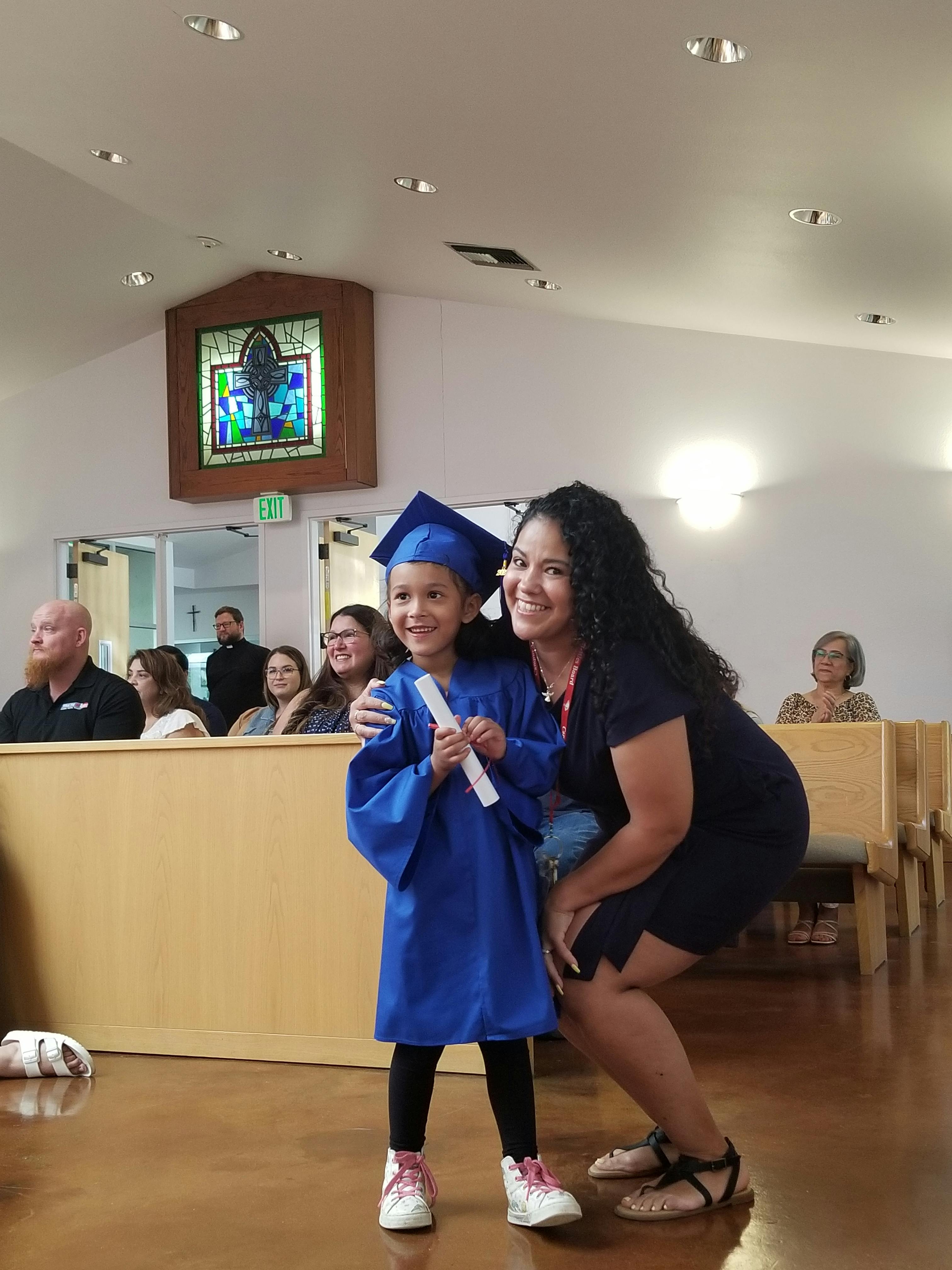 Child in blue graduation gown and cap holding diploma next to an adult