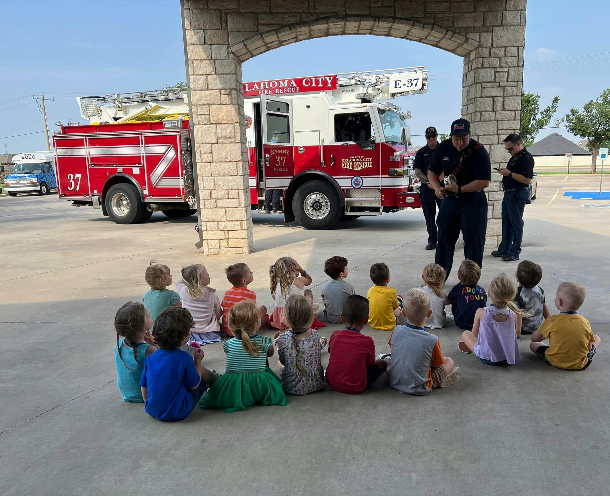 Children sitting on the ground listening to people in uniform near a fire truck
