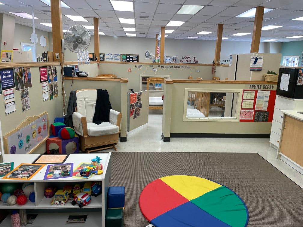 Classroom with child-sized furniture and a rocking chair, cribs visible through a window