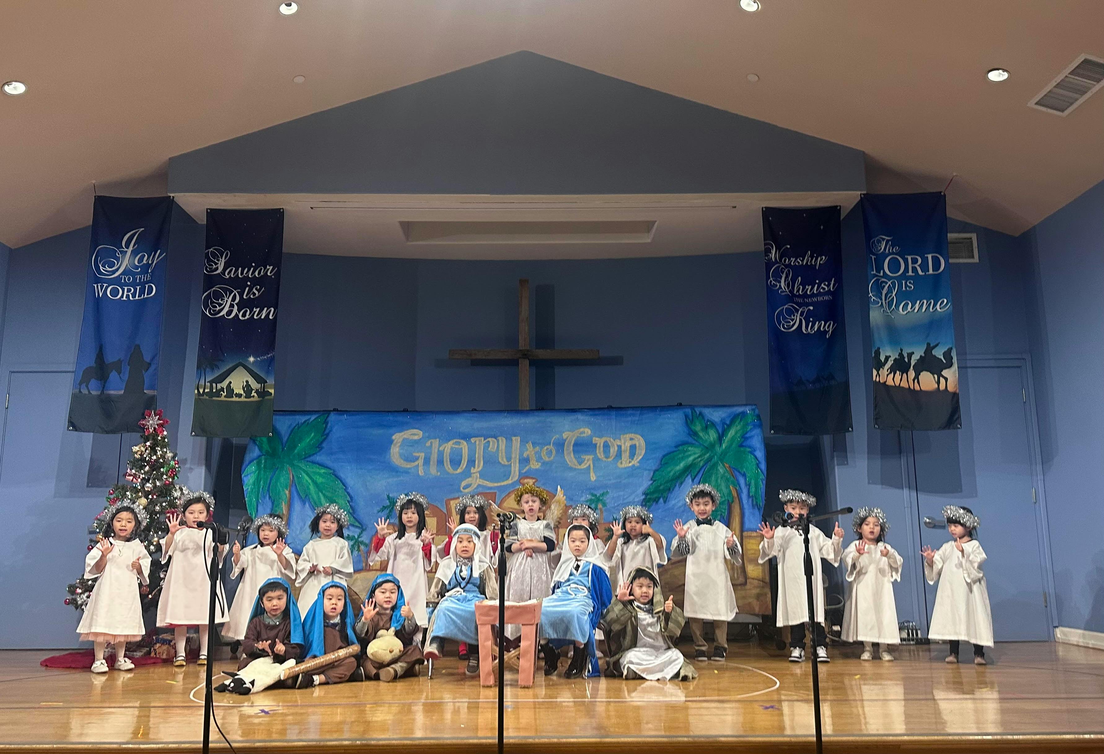 Group of children in costume performing on a stage under Glory to God banner