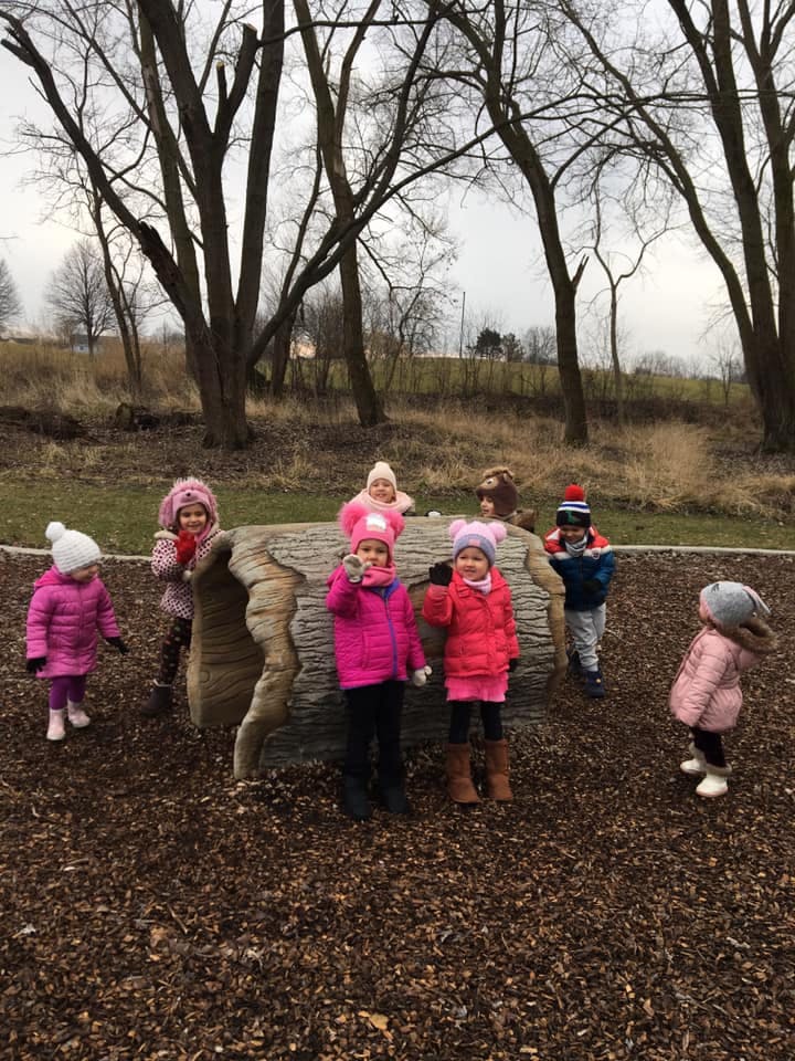 Children in winter coats and hats playing around a large log outdoors