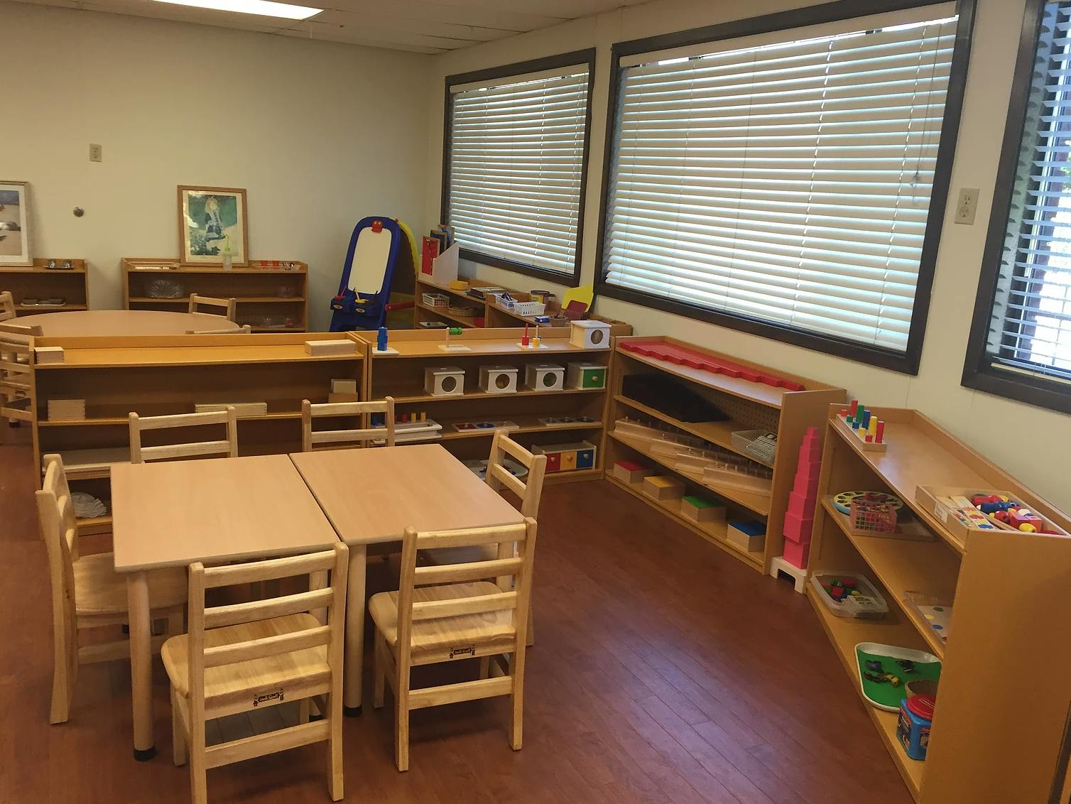 Empty classroom with small wooden tables and chairs, shelves with learning materials
