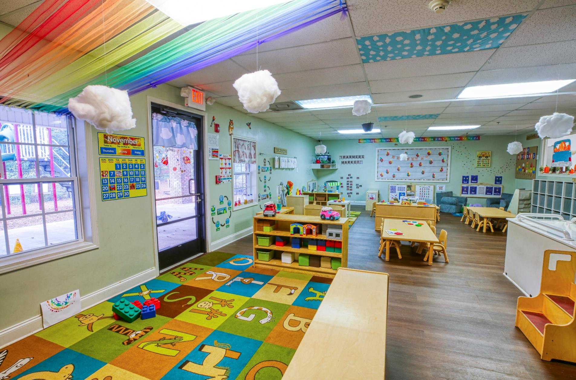 Colorful classroom with child-sized tables and chairs, a play rug, and shelves with a November calendar on the wall