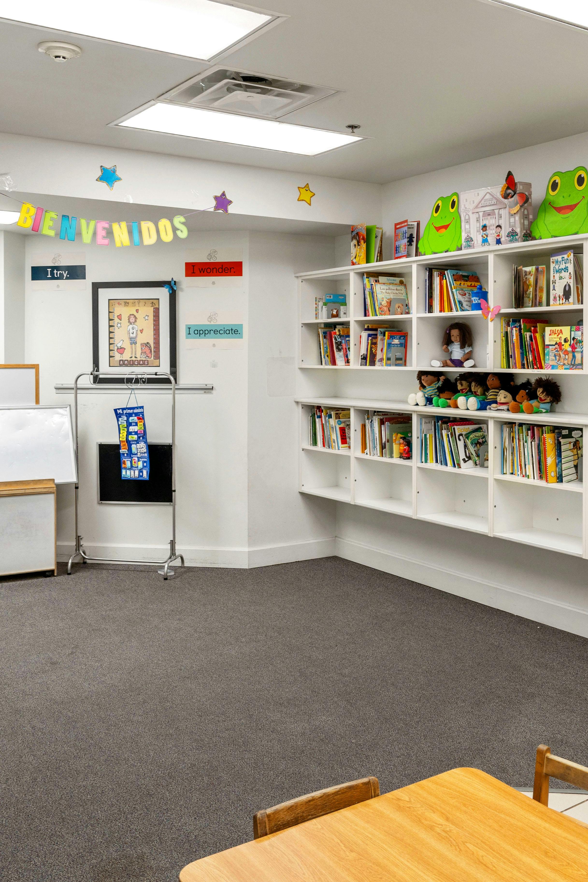Classroom with "Bienvenidos" sign, whiteboards, and bookshelves filled with books and toys