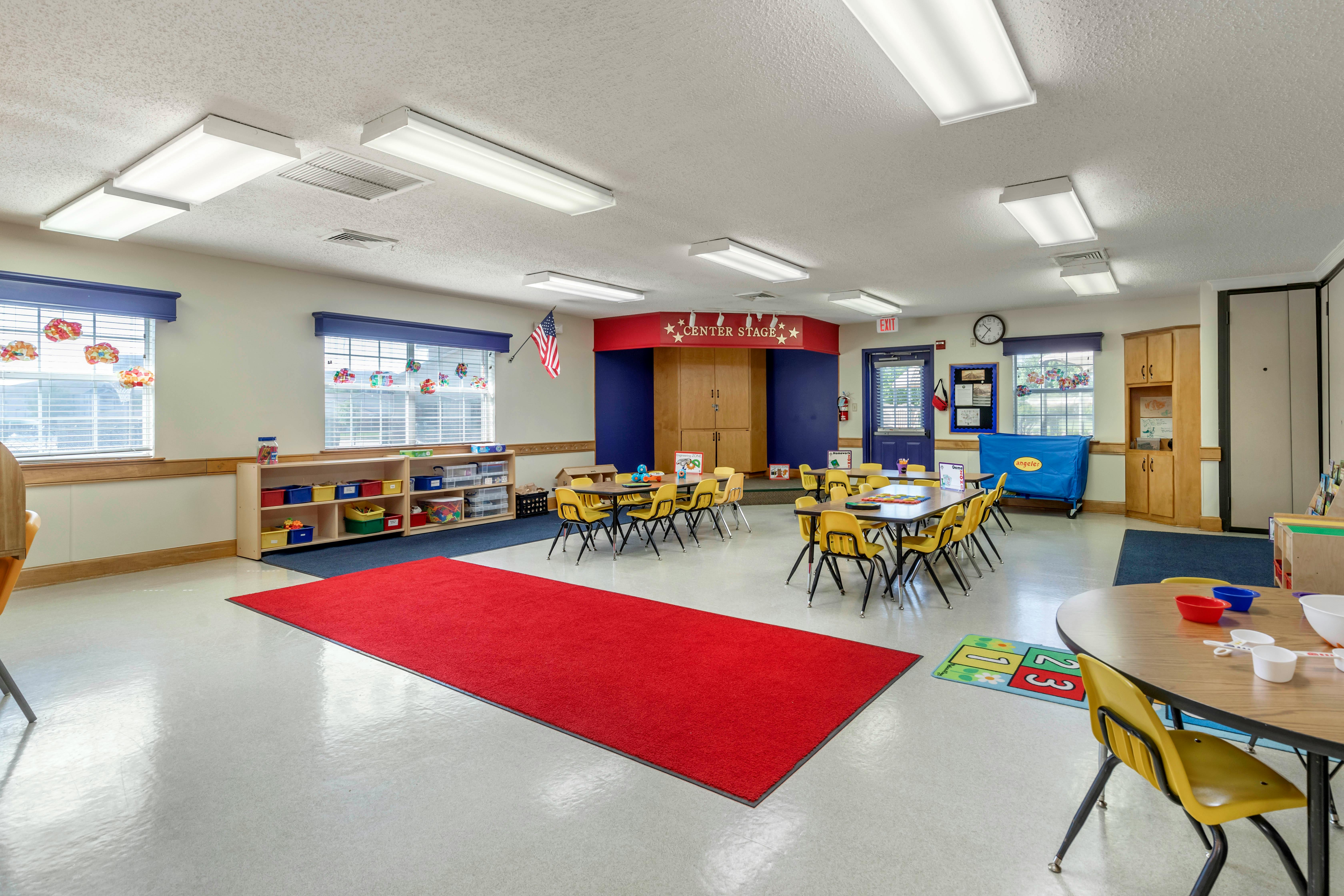 An empty classroom with yellow tables and chairs, a red carpet leading to a stage