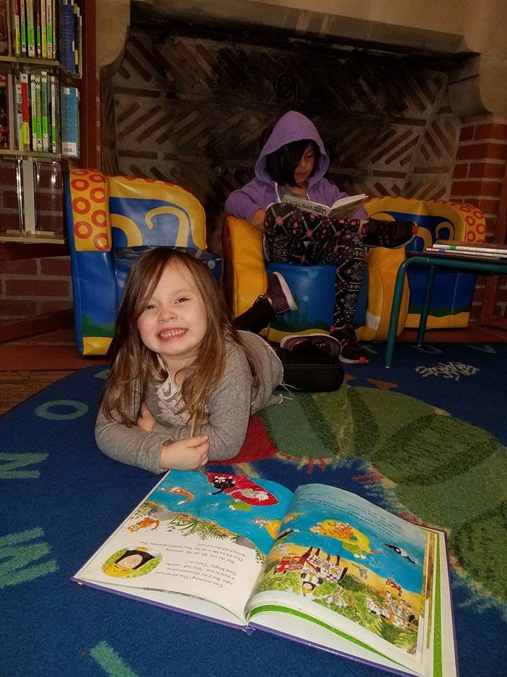 Child lying on floor with open book, person in background reading