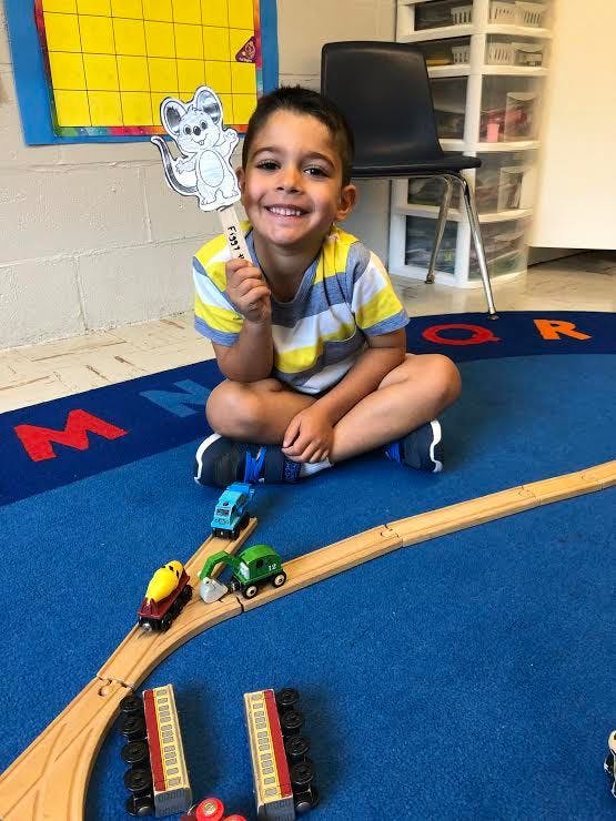 Child holding up a paper mouse puppet while sitting on a blue rug with toy trains