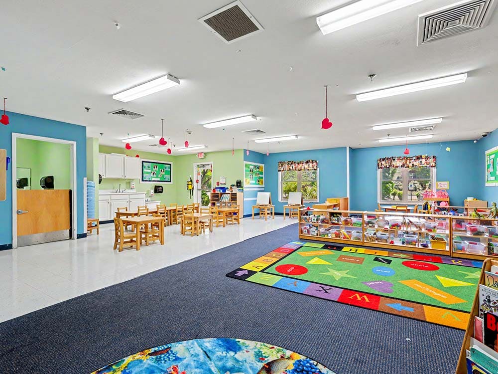 Colorful classroom with small tables and chairs, a play rug, and windows