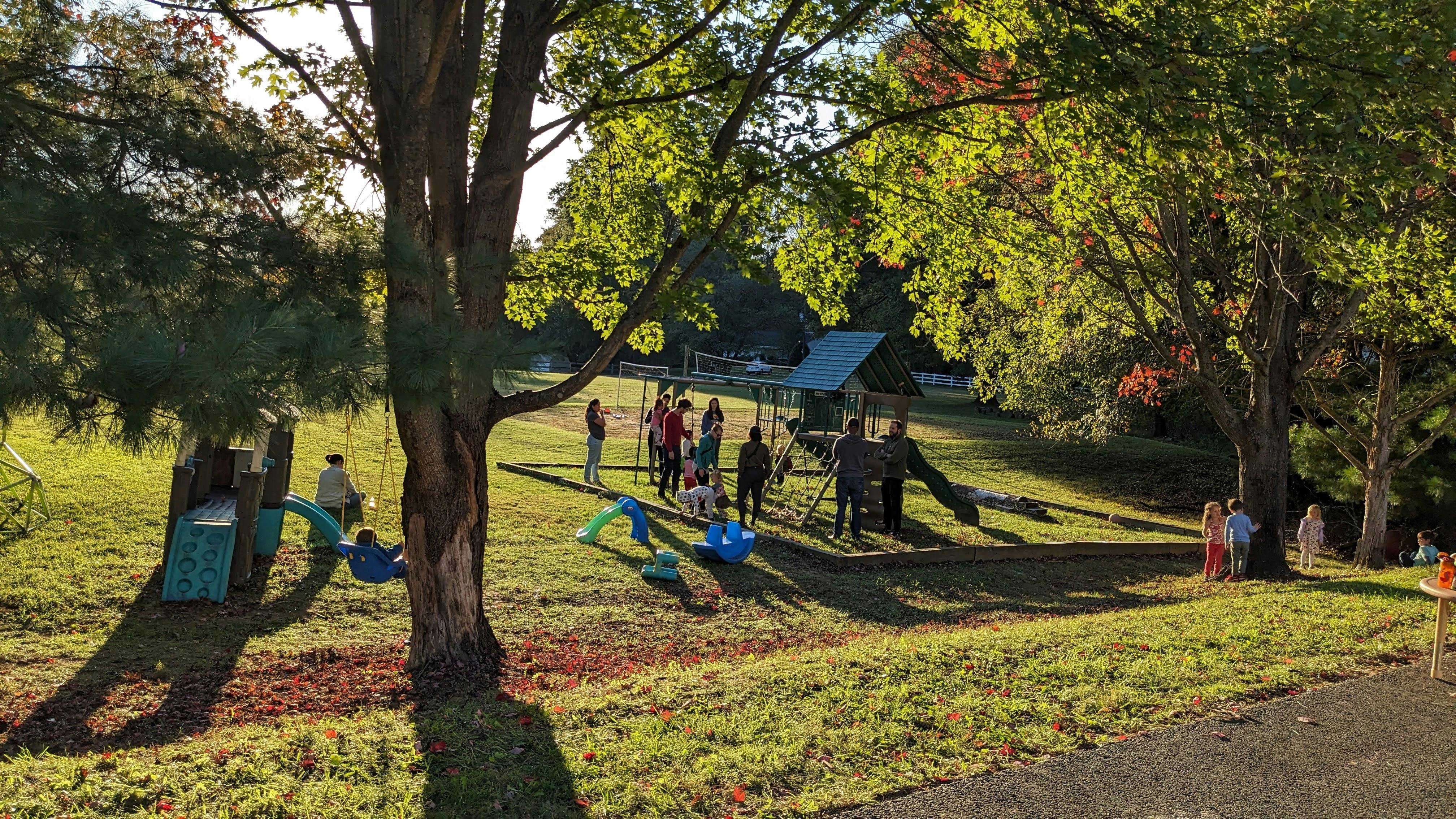People and children gathered around playground equipment under trees