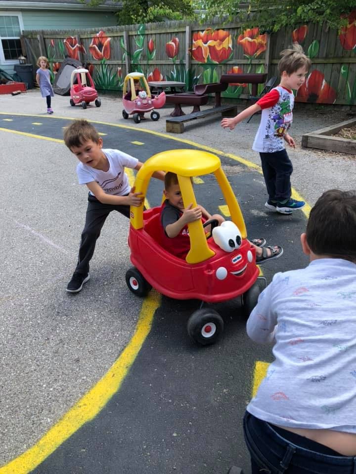 Child pushing another child in a toy car on a paved area