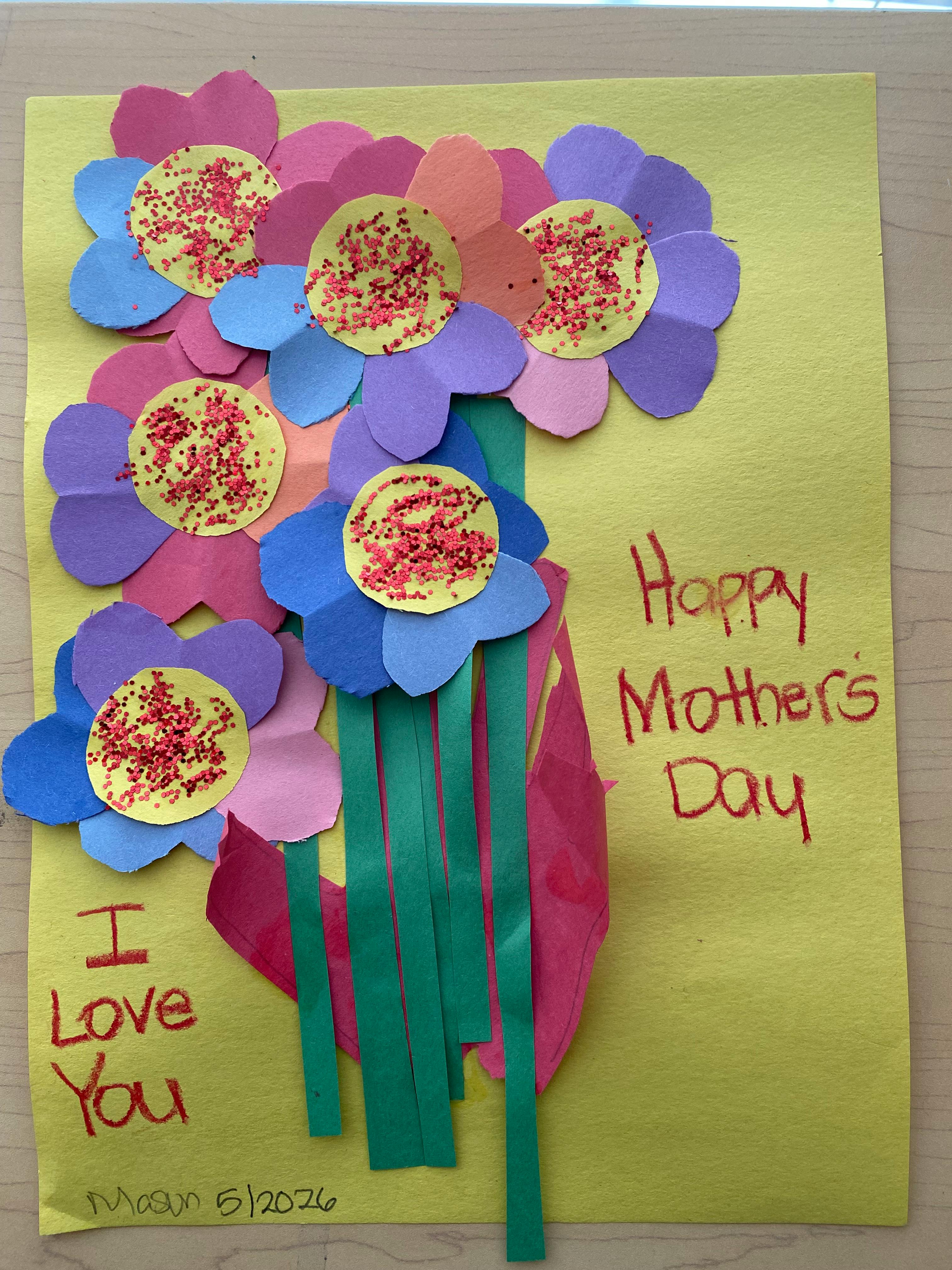 Child's craft project of colorful paper flowers with glitter and text "Happy Mother's Day"