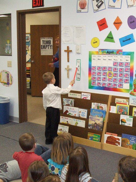 Child pointing at a calendar on a wall in a classroom with other children watching