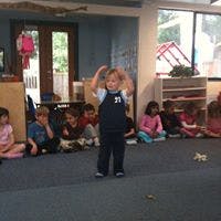 Child standing with hands on head, other children sitting on floor