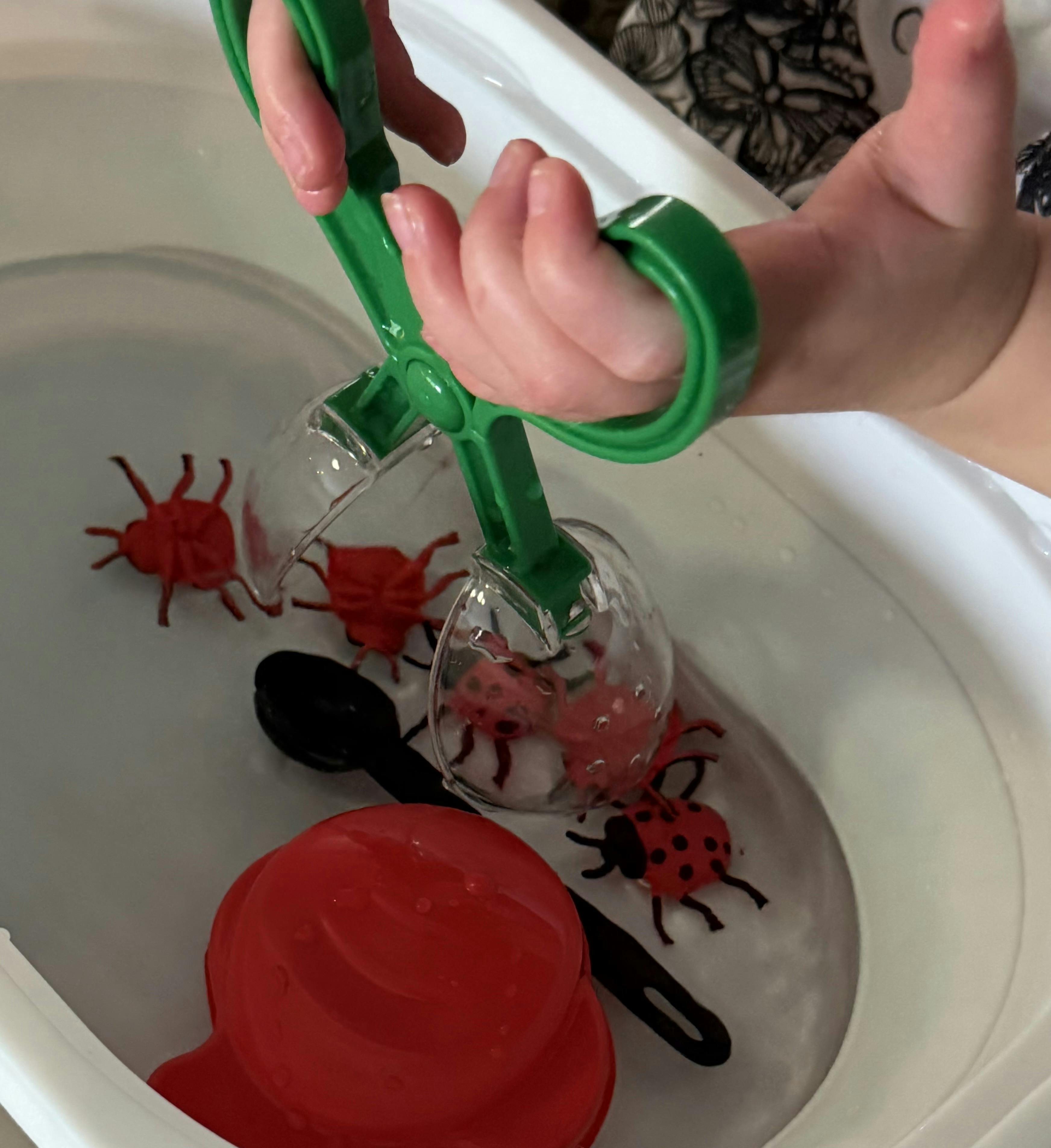 Child using plastic scoopers to pick up toy insects in a water bin