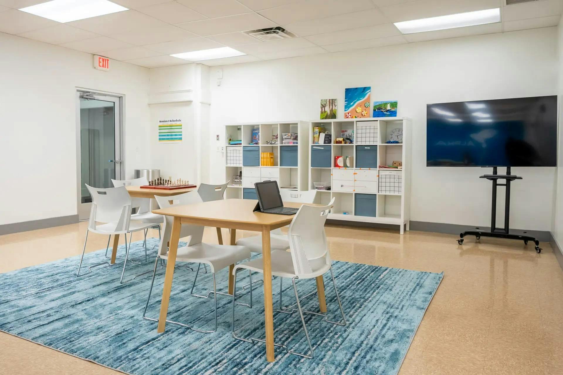 Classroom with tables, chairs, a blue rug, shelves, and a large TV