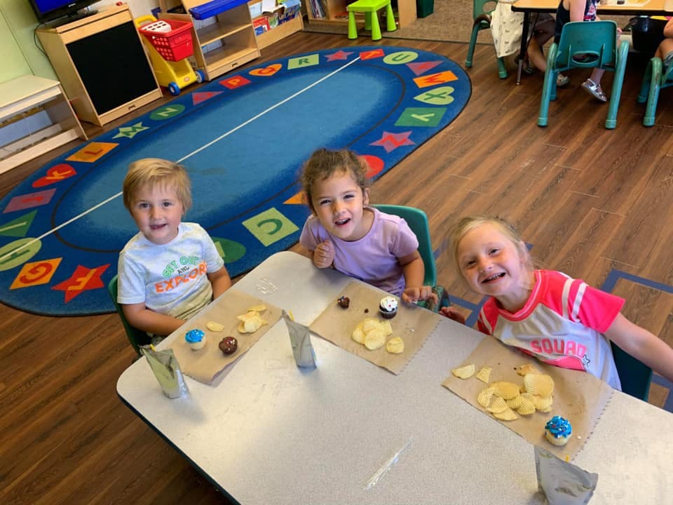 Three children sitting at a table with snacks in a classroom