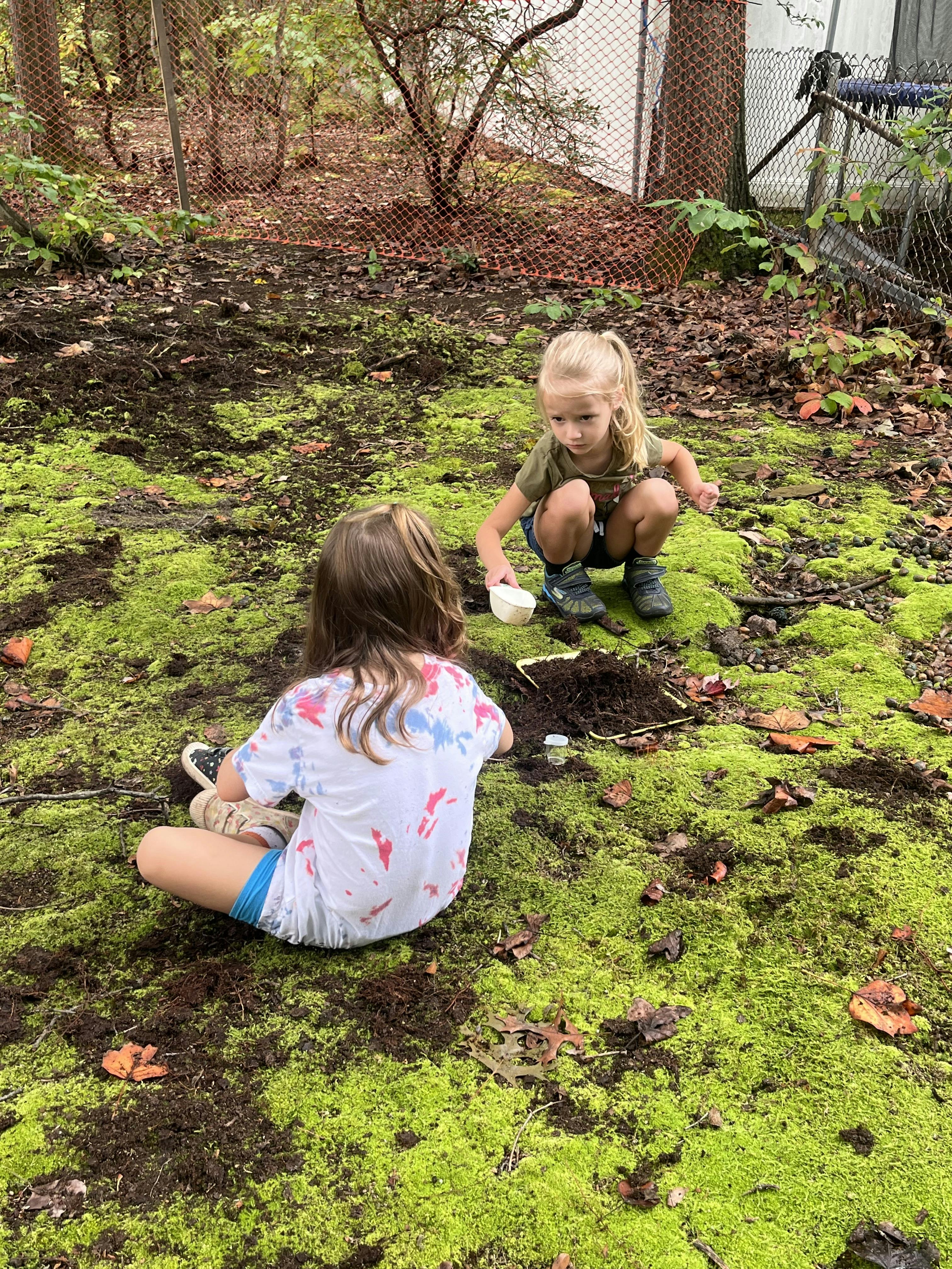 Two children playing in dirt and moss, one with a white scoop