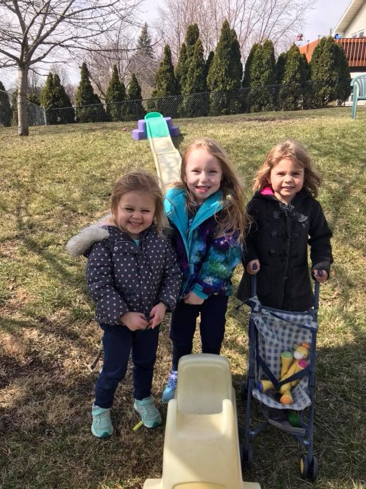 Three children standing outdoors near a small slide and a toy stroller