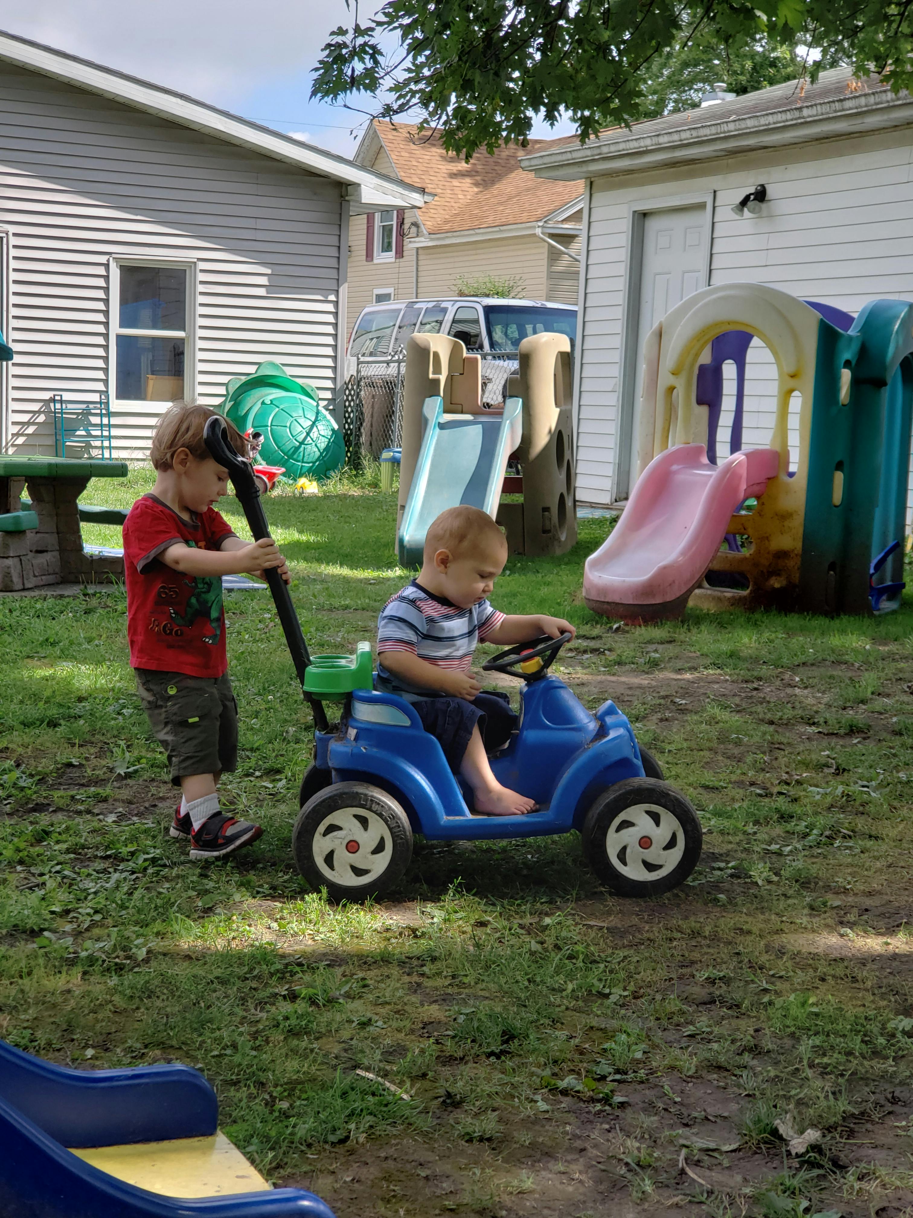Child pushing another child in a toy car in a yard