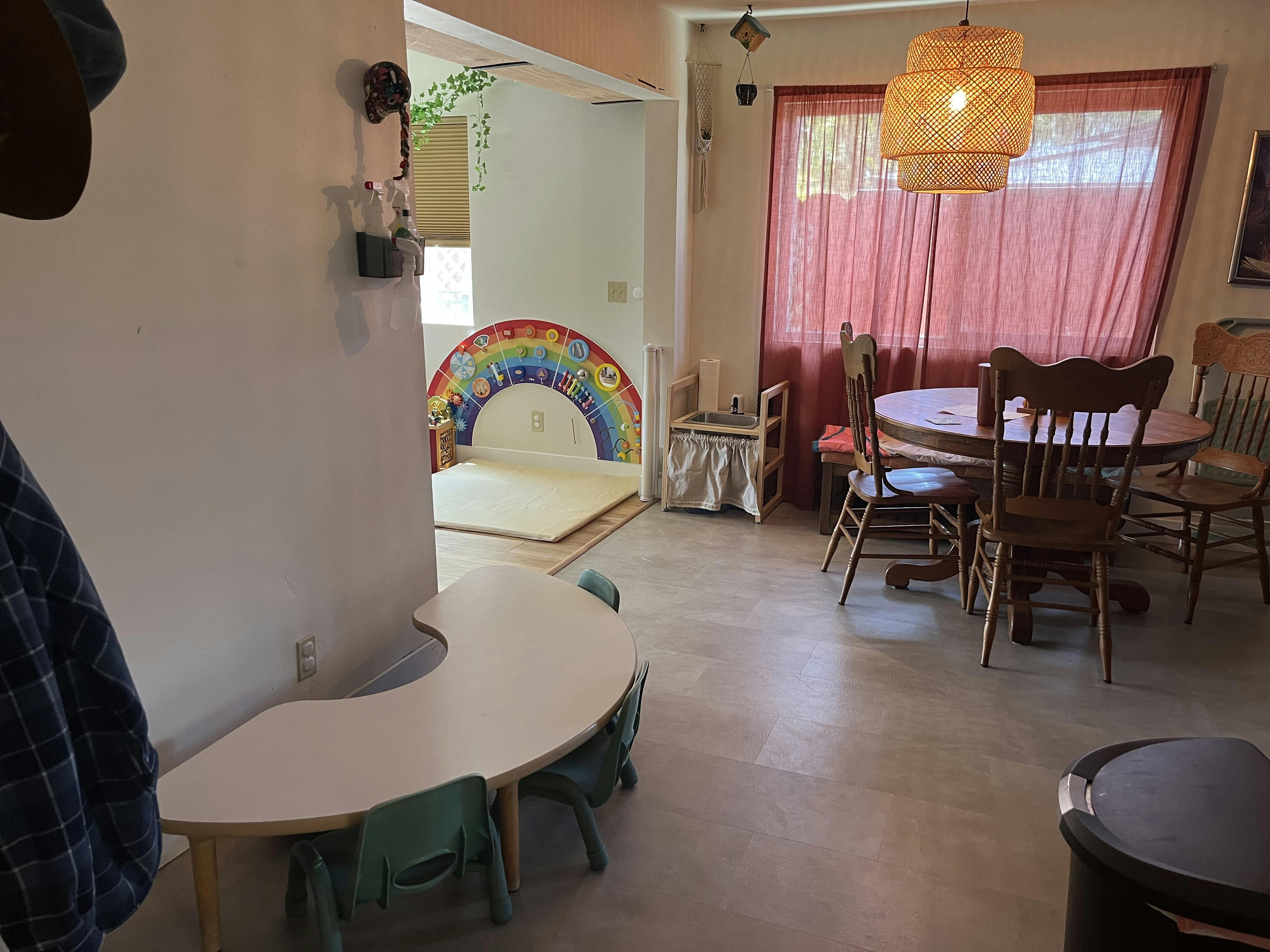 A room with a small table and chairs, a dining table, and a rainbow-colored play arch