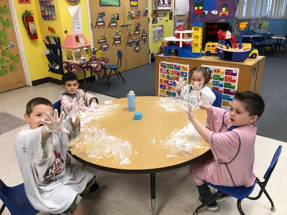 Three children playing with white foam at a table in a classroom