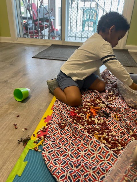 Child kneeling on a mat with beans, reaching into a container
