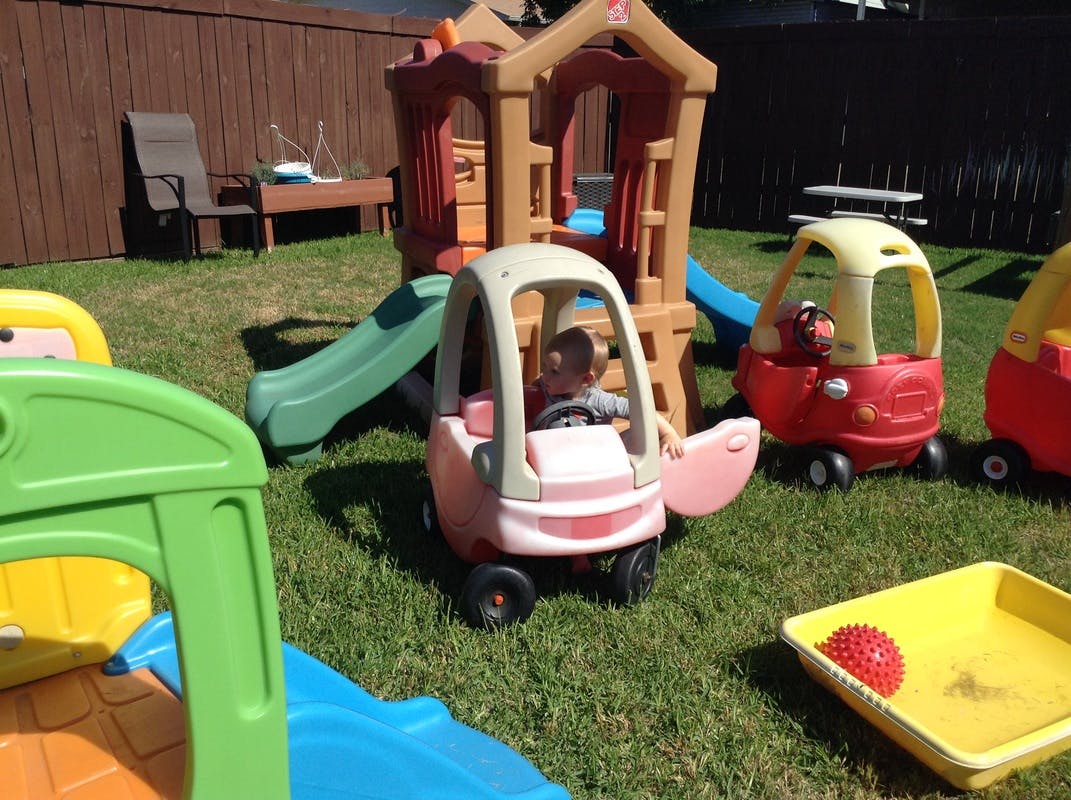 Child in a toy car on a grassy lawn with other play equipment