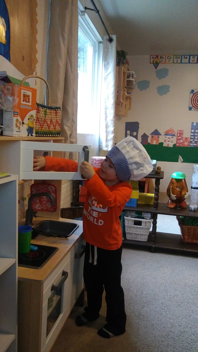 Child in a chef hat playing with a toy microwave in a play kitchen