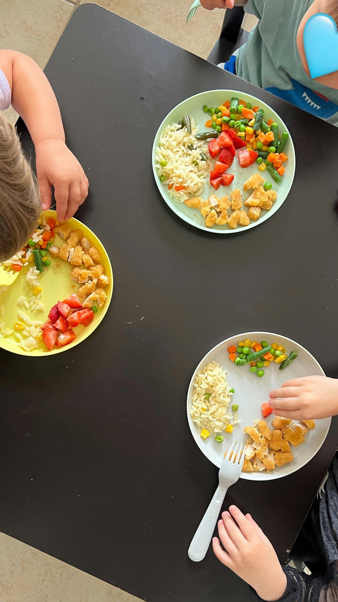 Multiple children eating plates of chicken, rice, strawberries, and mixed vegetables