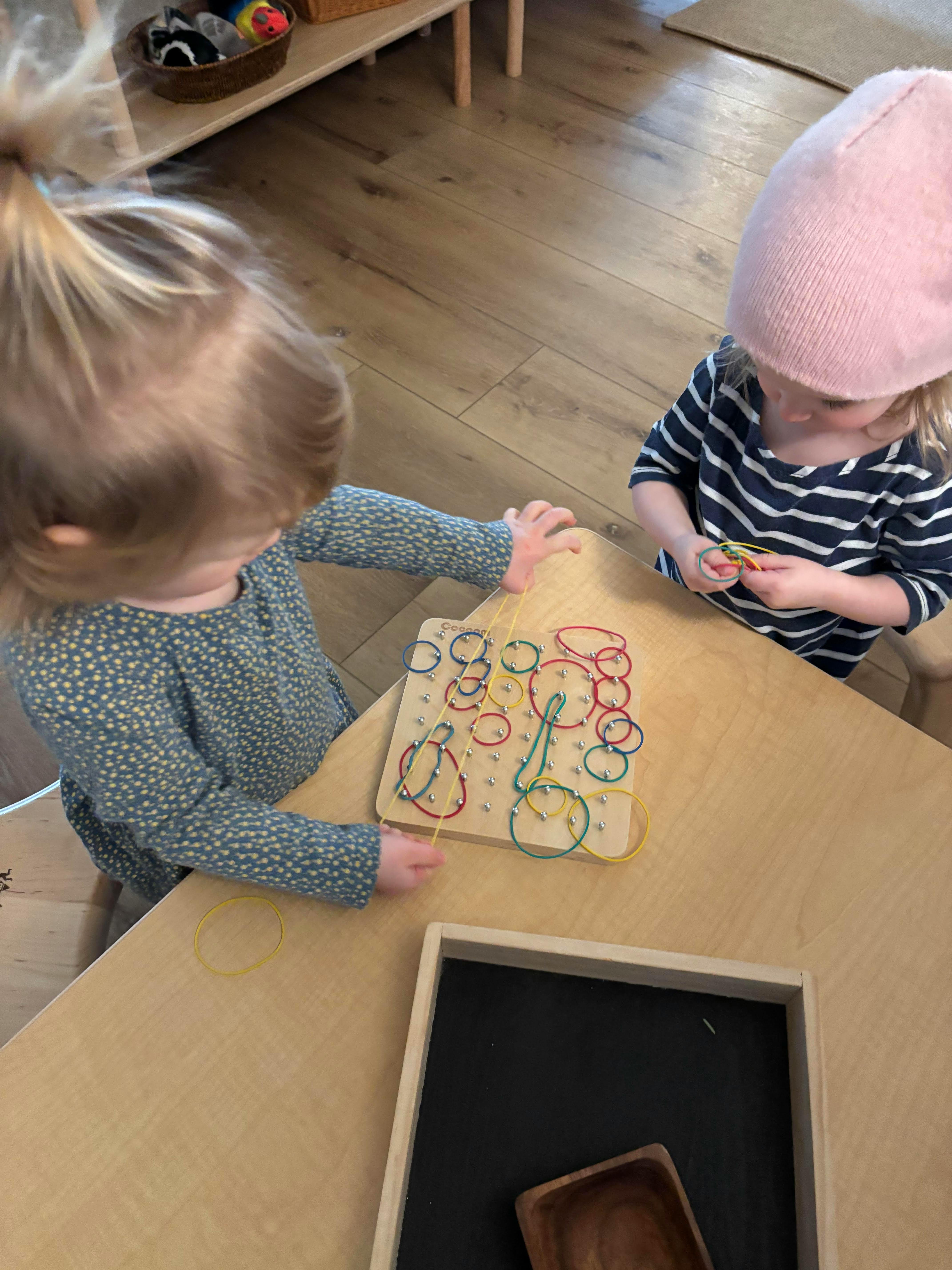 Two children playing with colorful rubber bands on a pegboard on a wooden table