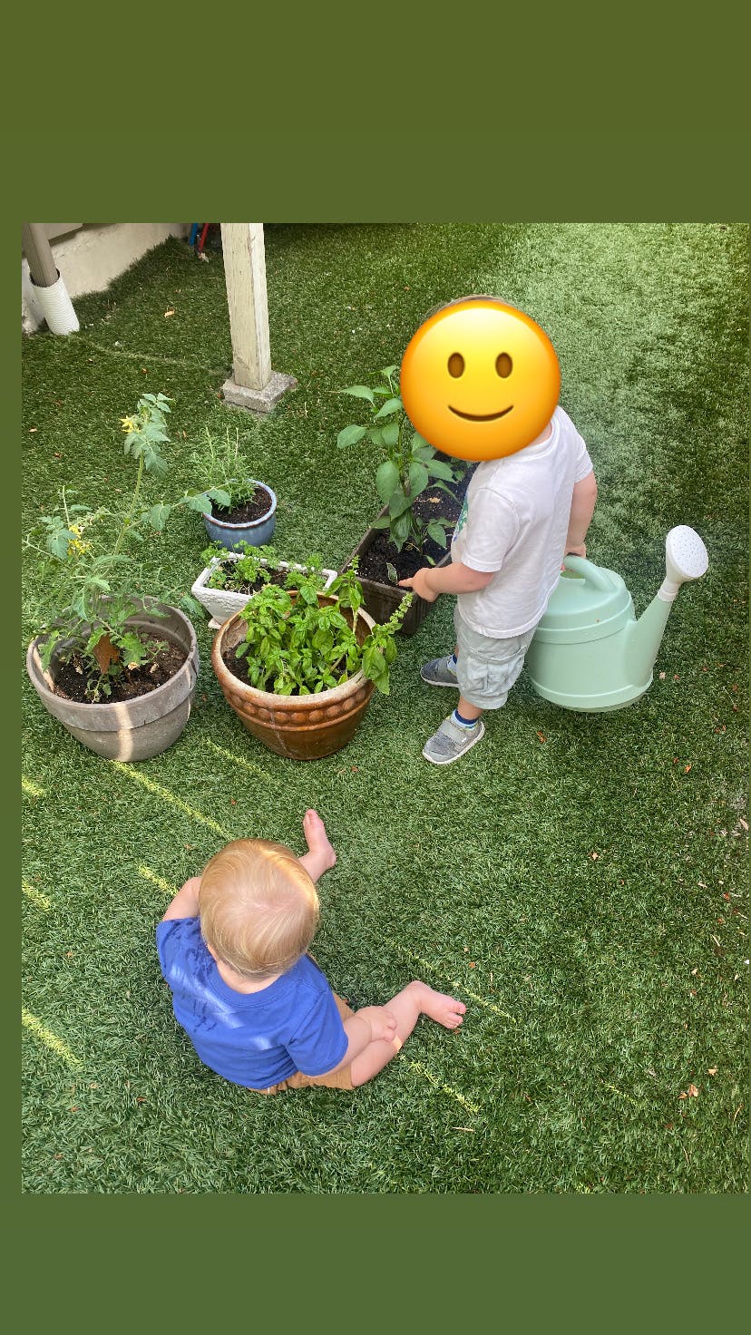Child watering plants in pots on grass, another child sitting nearby