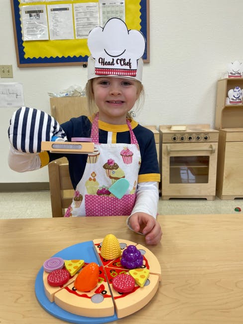 Child wearing a chef hat and apron, holding a toy pizza cutter over a toy pizza