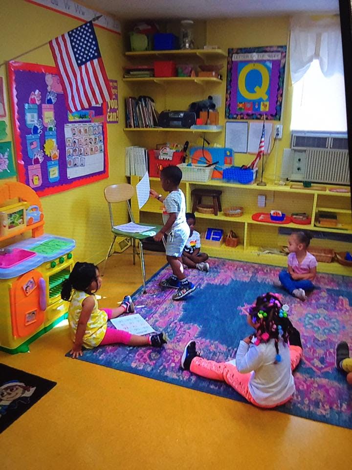 Children in a classroom, some sitting on a rug, one standing