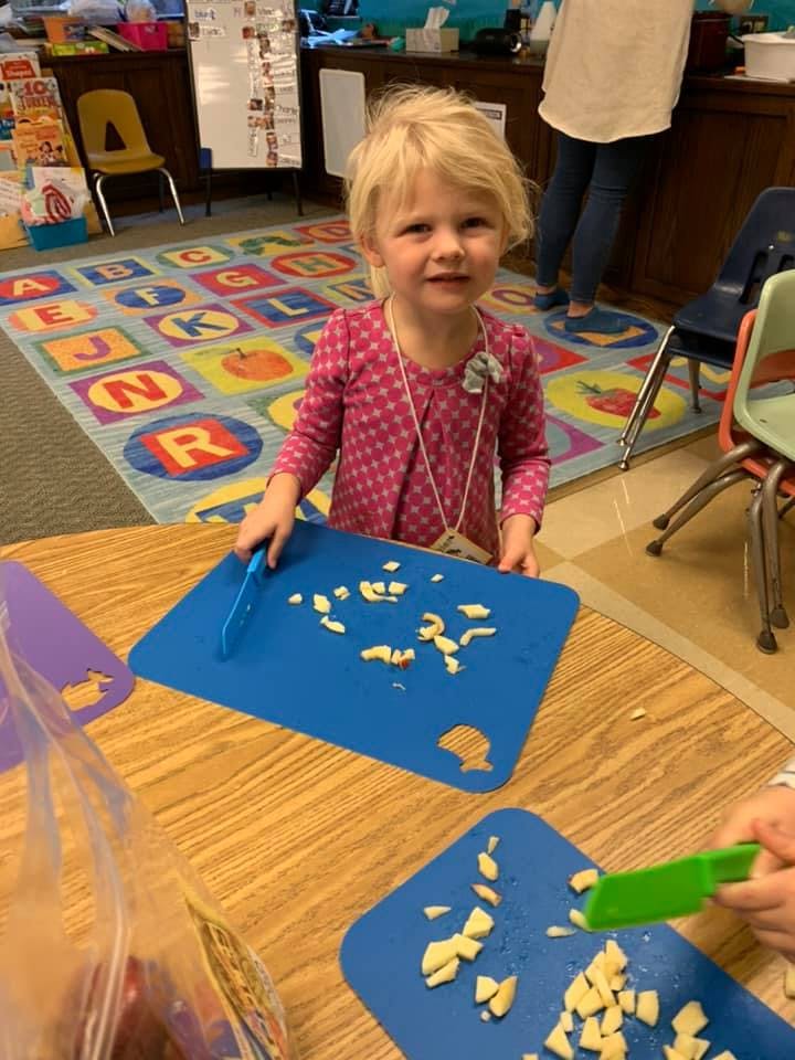 Child cutting food on a blue cutting board in a classroom