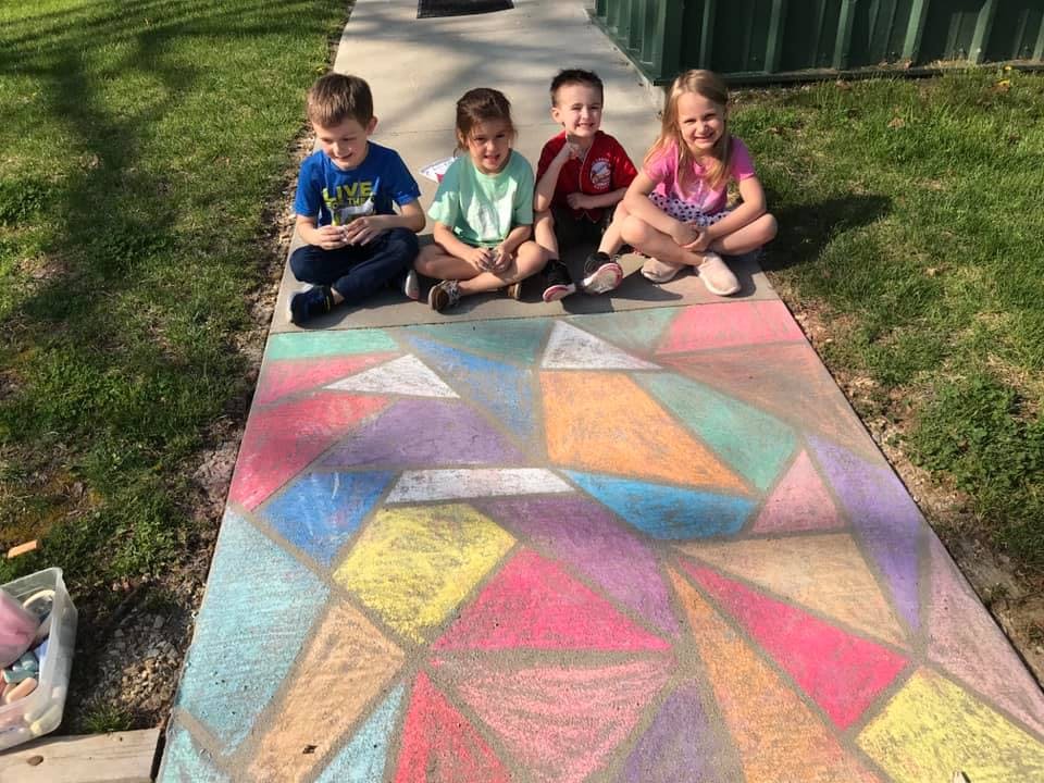Four children sitting on a sidewalk next to a colorful chalk drawing