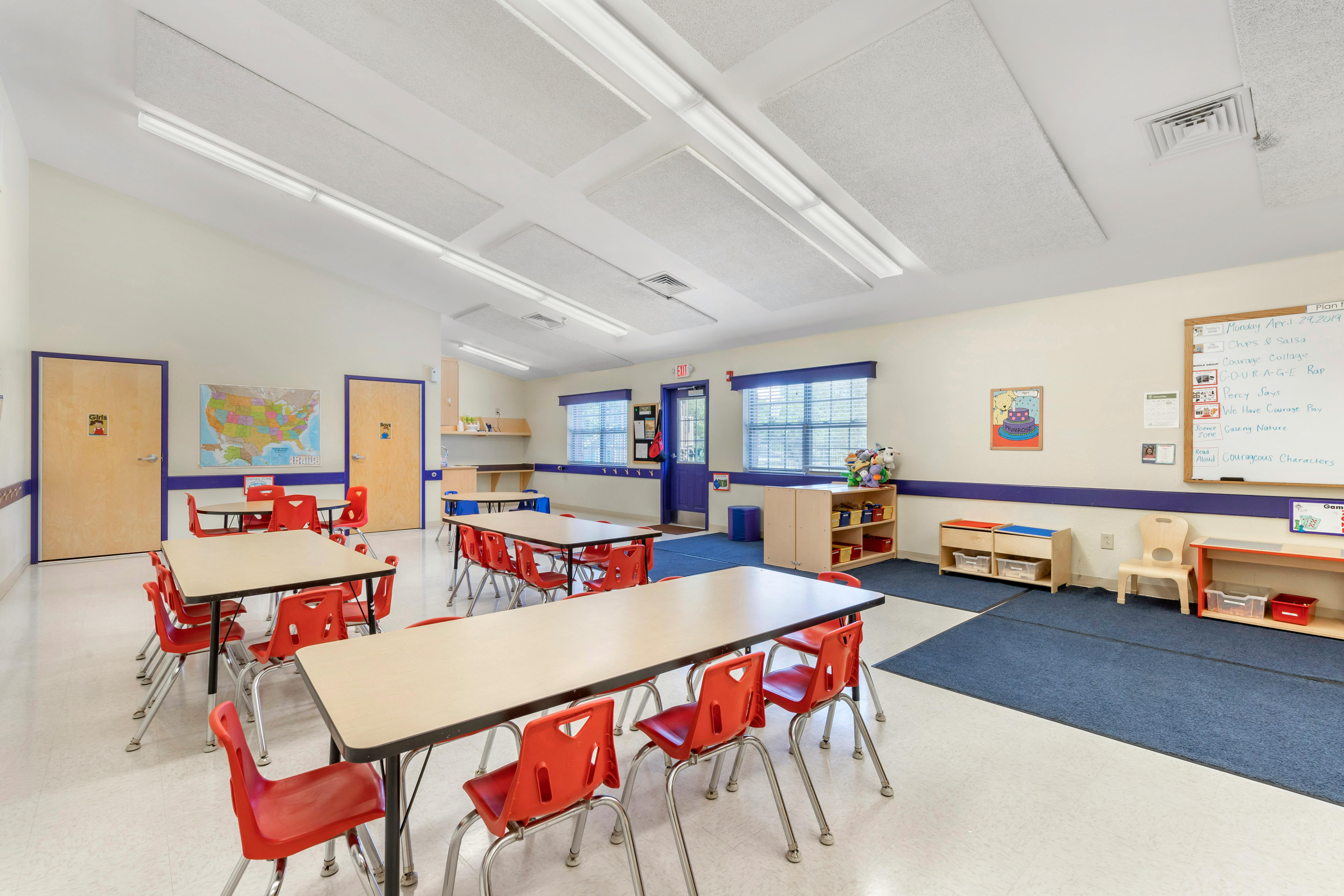 Empty classroom with small tables and red chairs, whiteboard with text, maps on tiled and carpeted floor