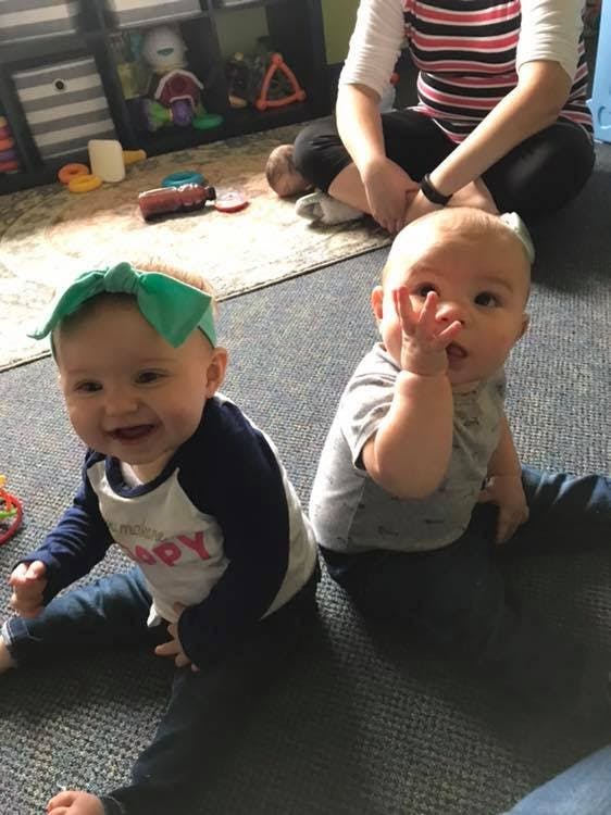 Two children sitting on a rug, one smiling and one with hand near face