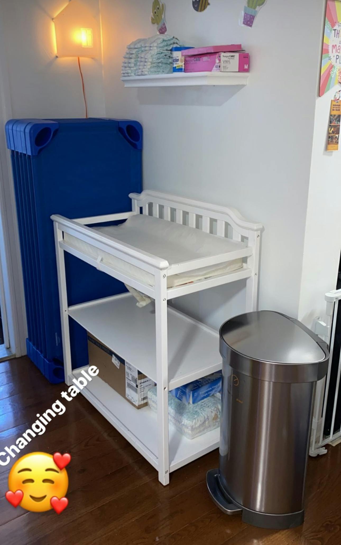 Changing table with shelves next to a blue cot and a trash can