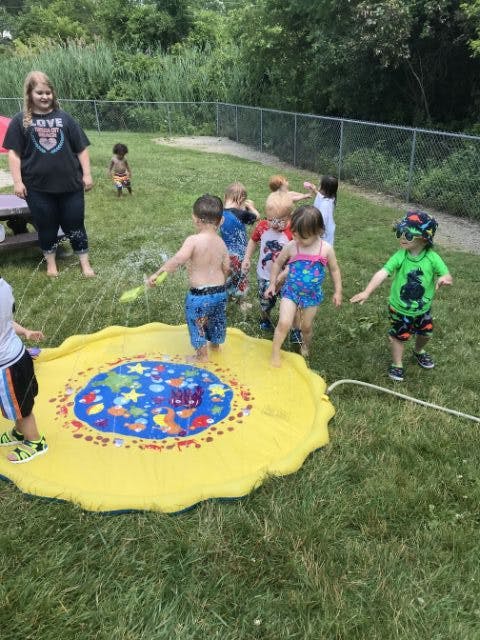 Person and children playing with a yellow splash pad and water in a grassy area