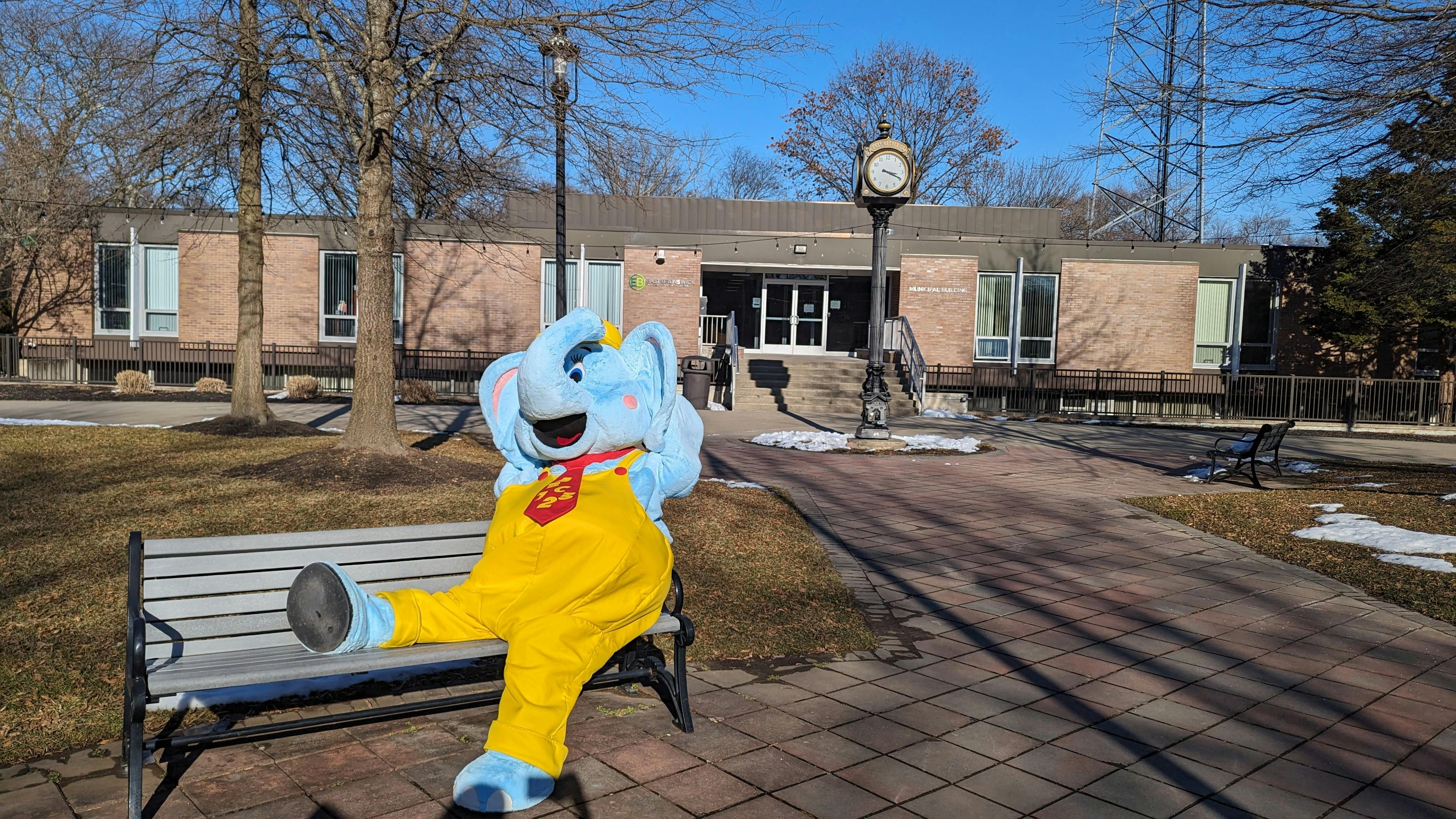 Mascot in blue and yellow overalls sitting on a park bench