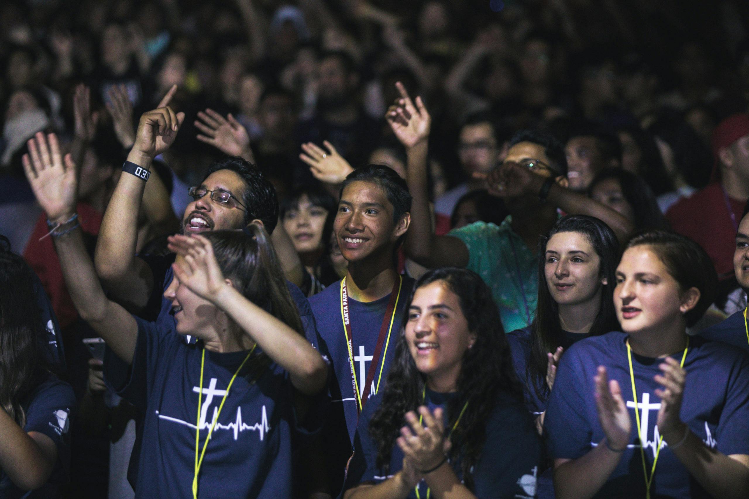 Group of people in dark t-shirts with a cross design, many raising their hands