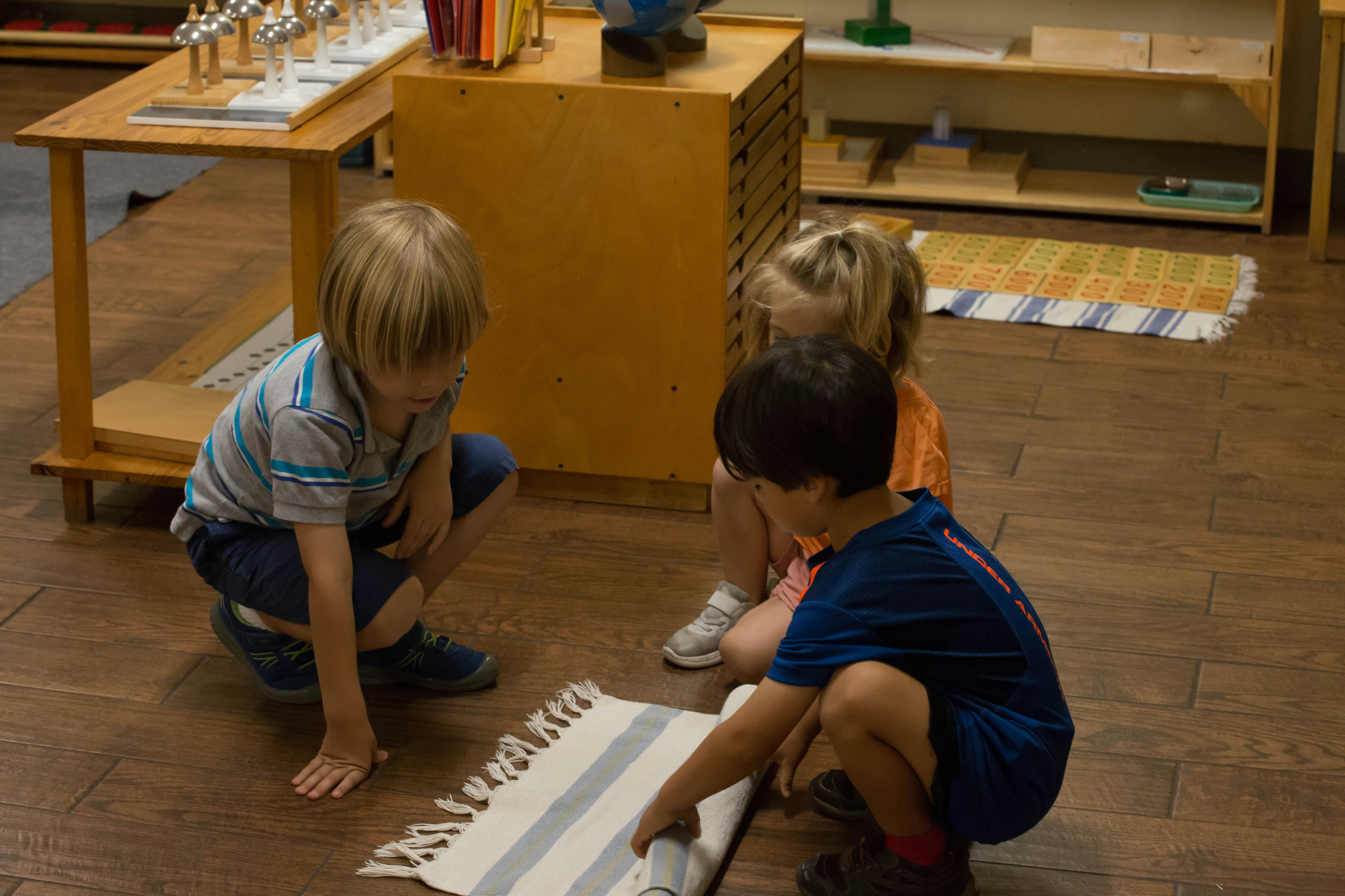 Three children crouching on a wooden floor, one rolling up a small rug