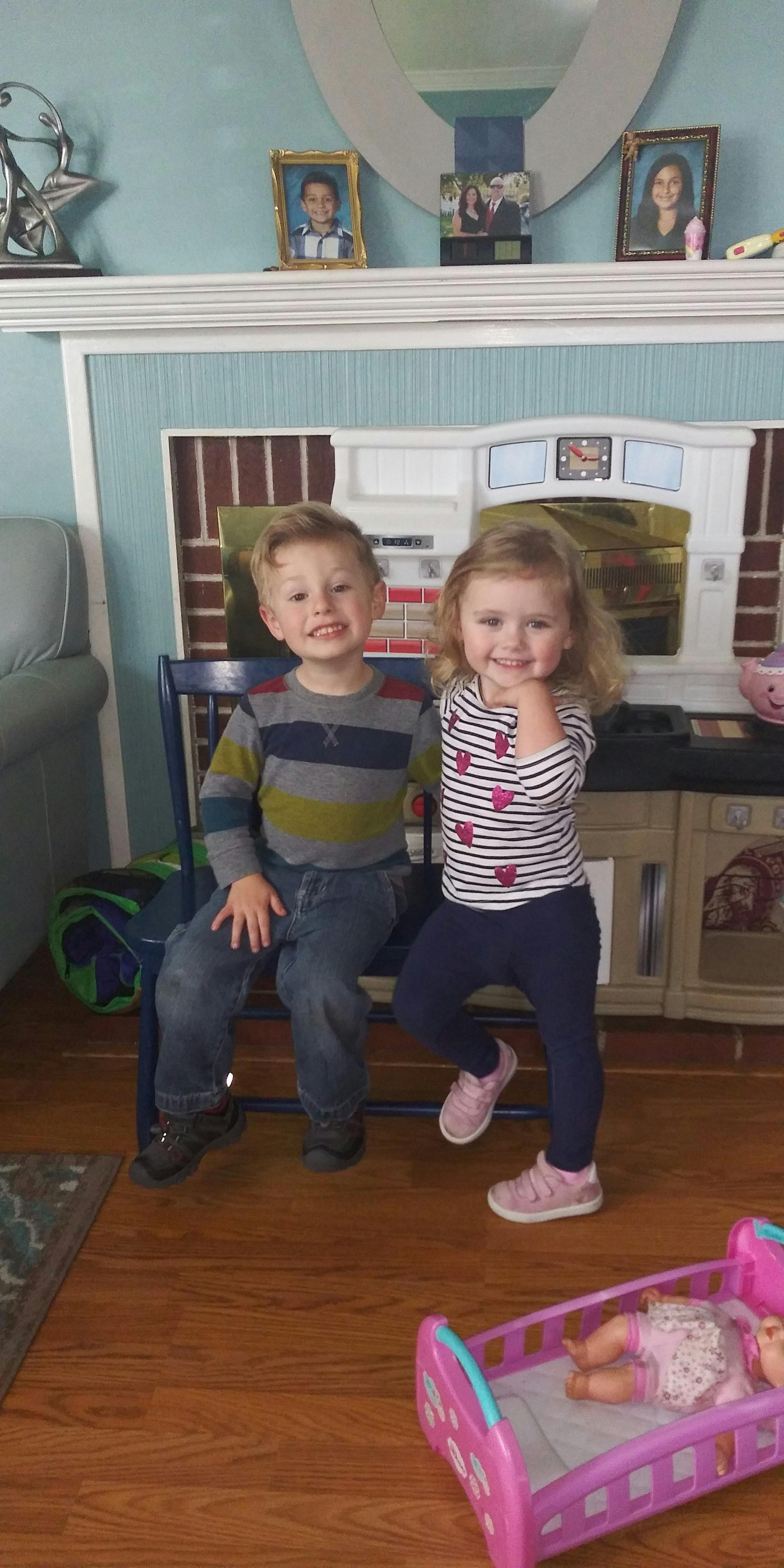 Two children smiling, one sitting on a chair, one standing