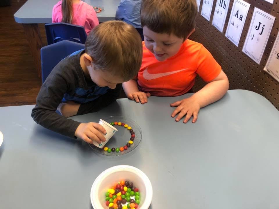 Two children at a table with candy, one pouring water onto a plate