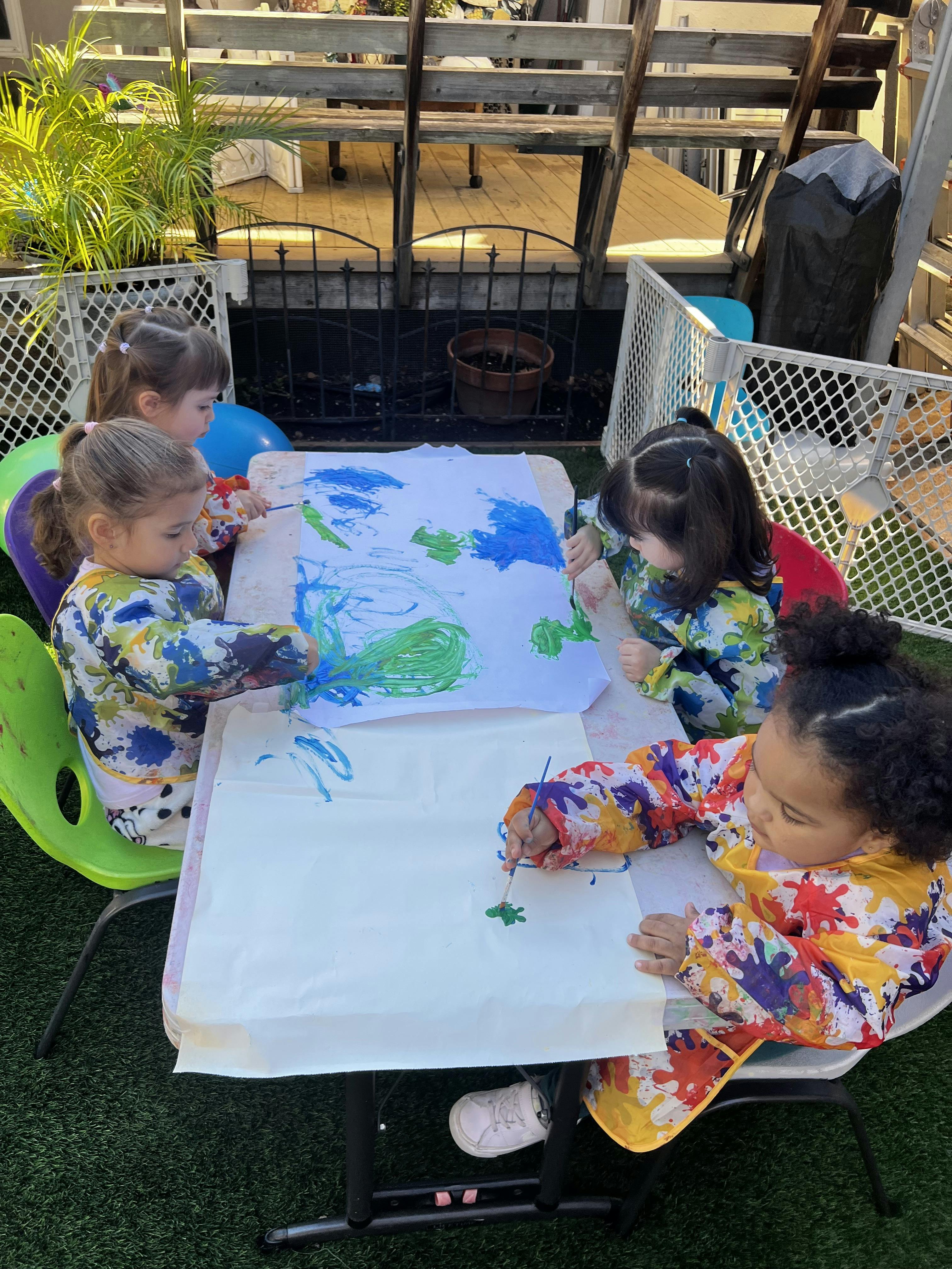 Four children in smocks painting at a table outdoors