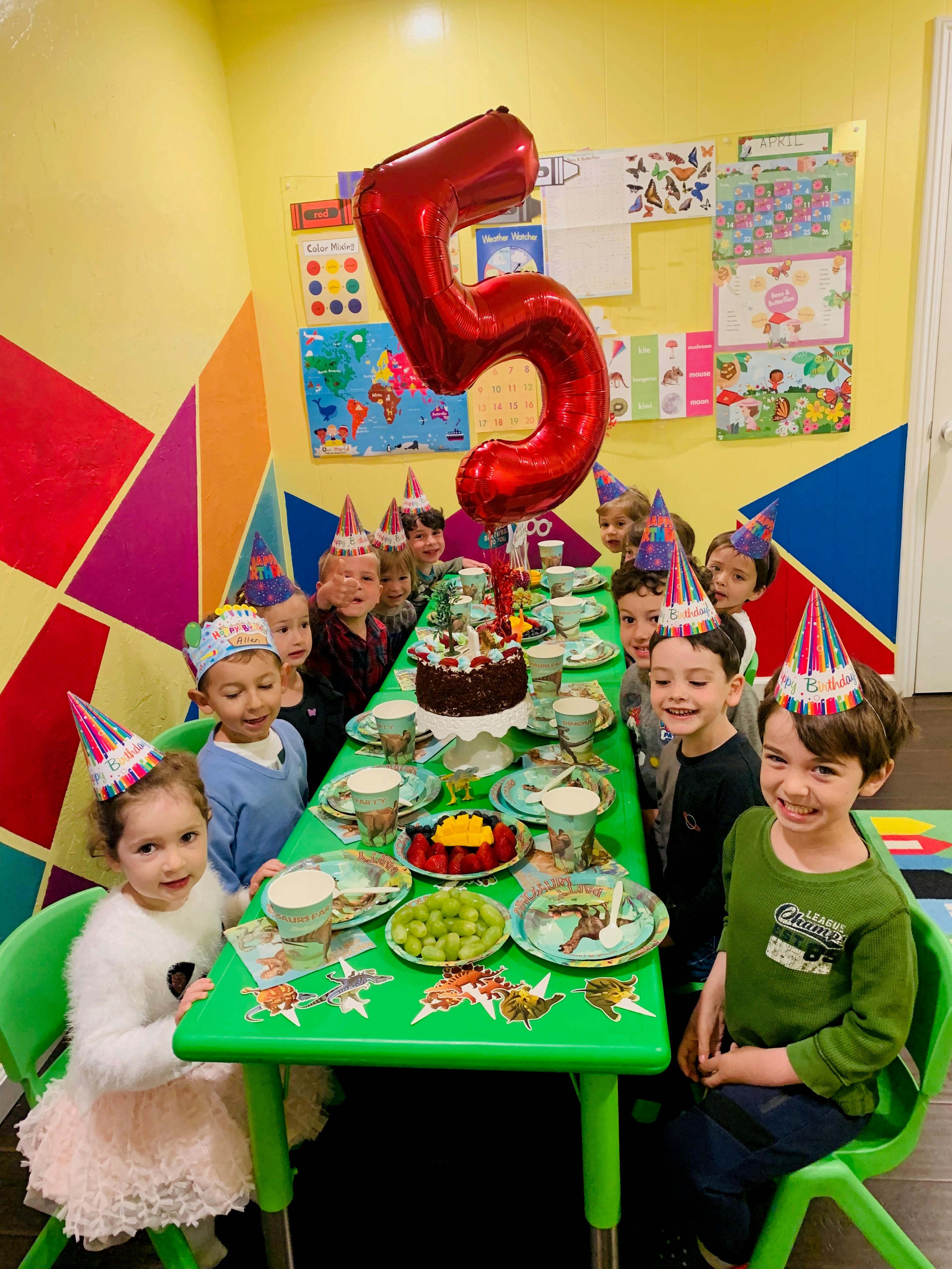 Children in party hats sitting around a table with a birthday cake and a large number 5 balloon