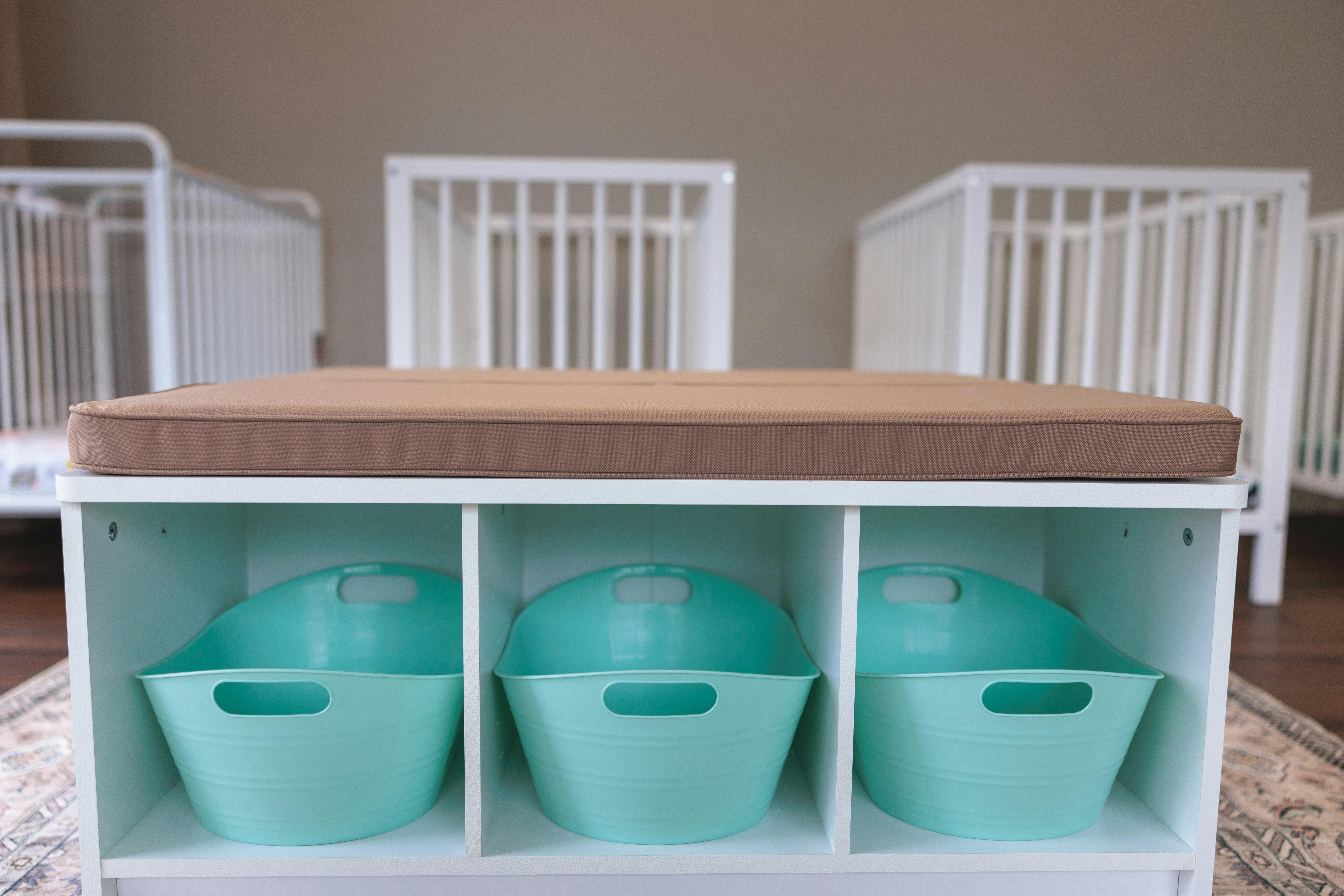 Storage unit with three teal bins and a brown cushion, cribs in background
