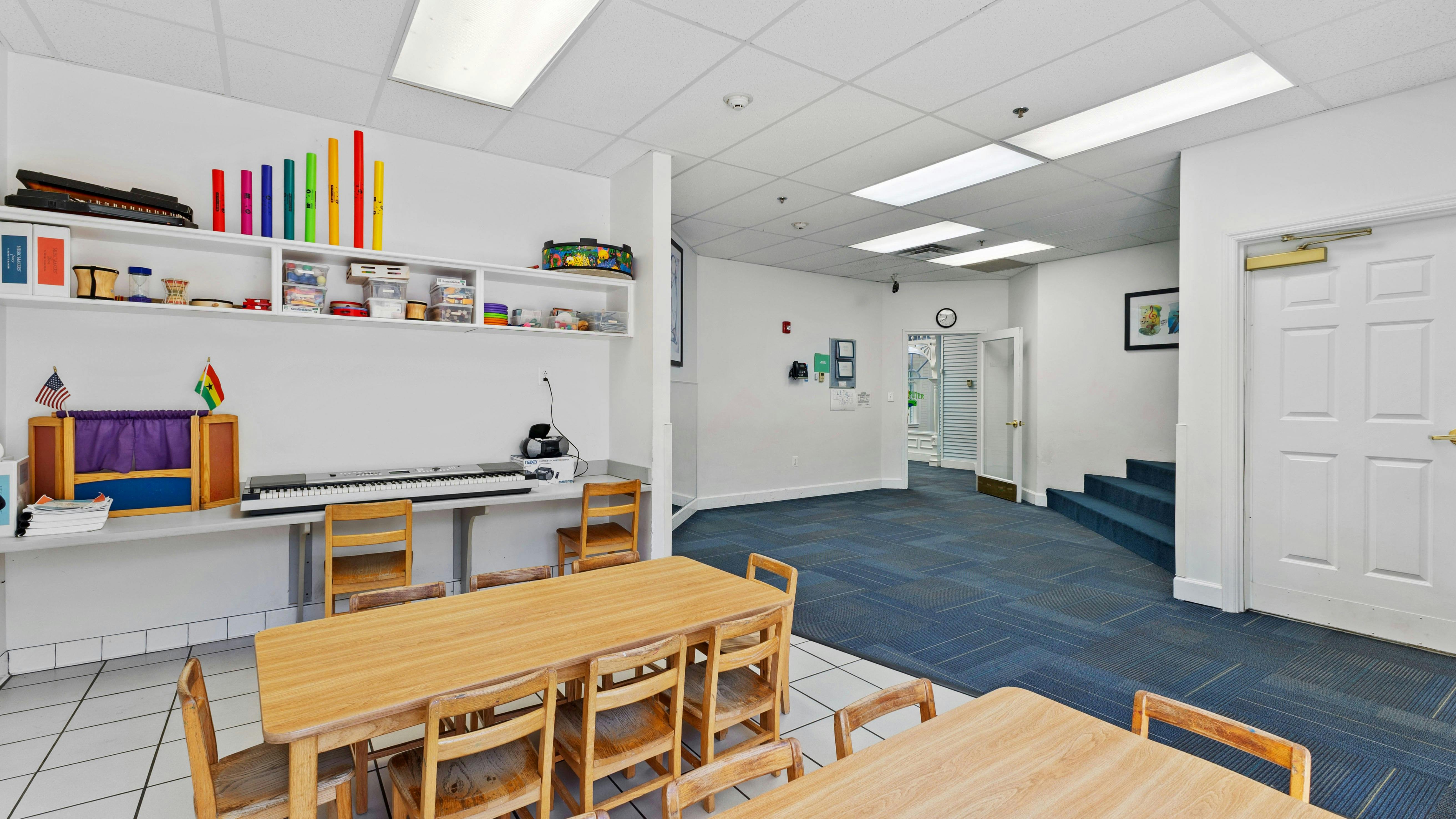 Empty classroom with small wooden tables and chairs, shelves with musical instruments, and a keyboard