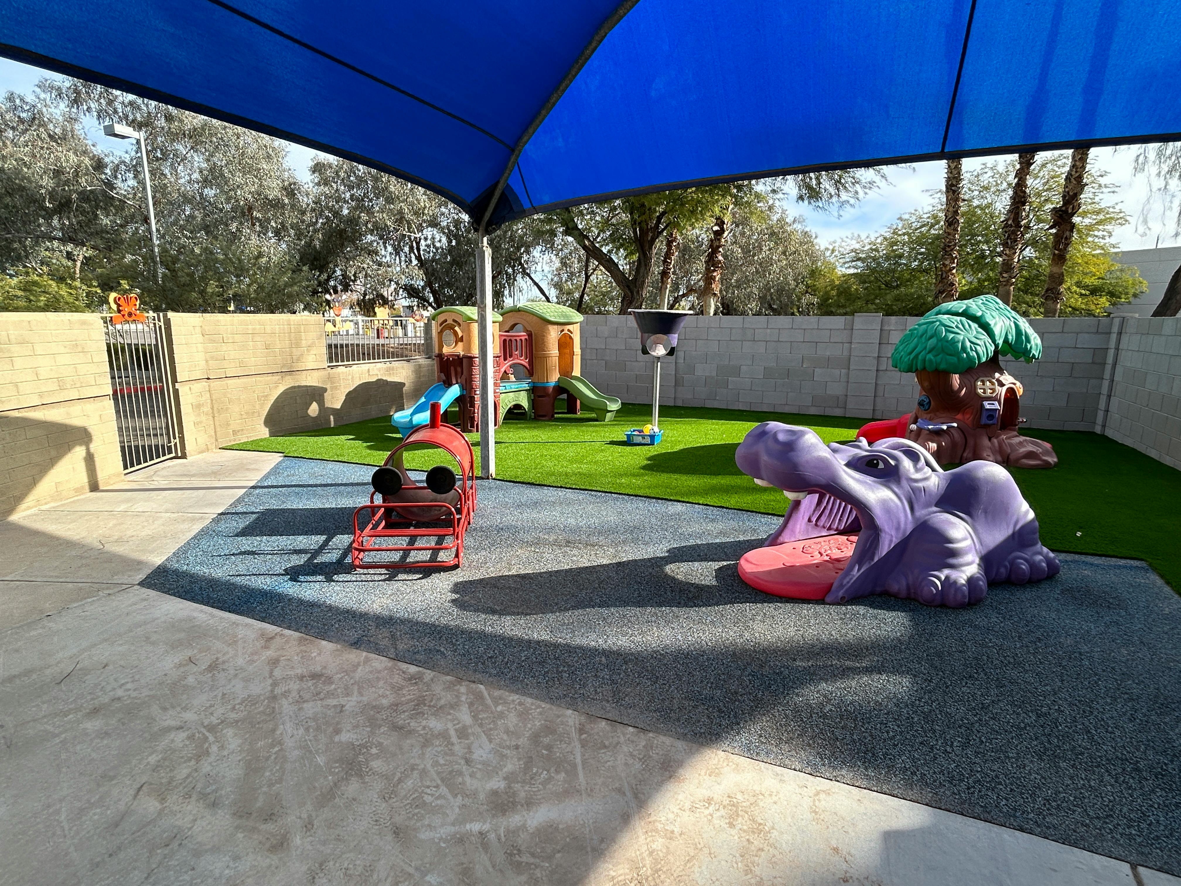 Outdoor playground with a blue shade canopy, play structures, and artificial turf