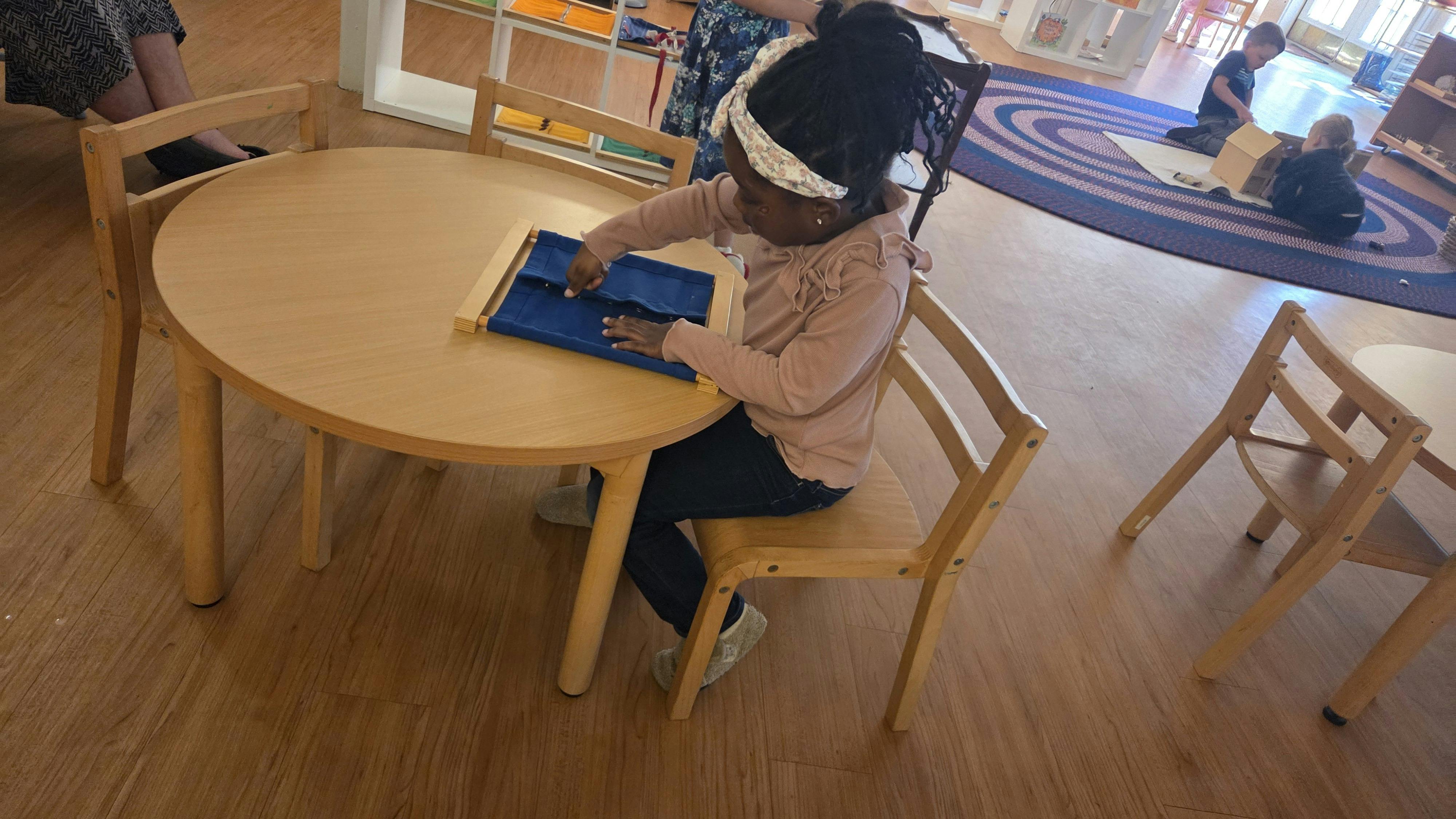 Child using a Montessori dressing frame at a table in a classroom